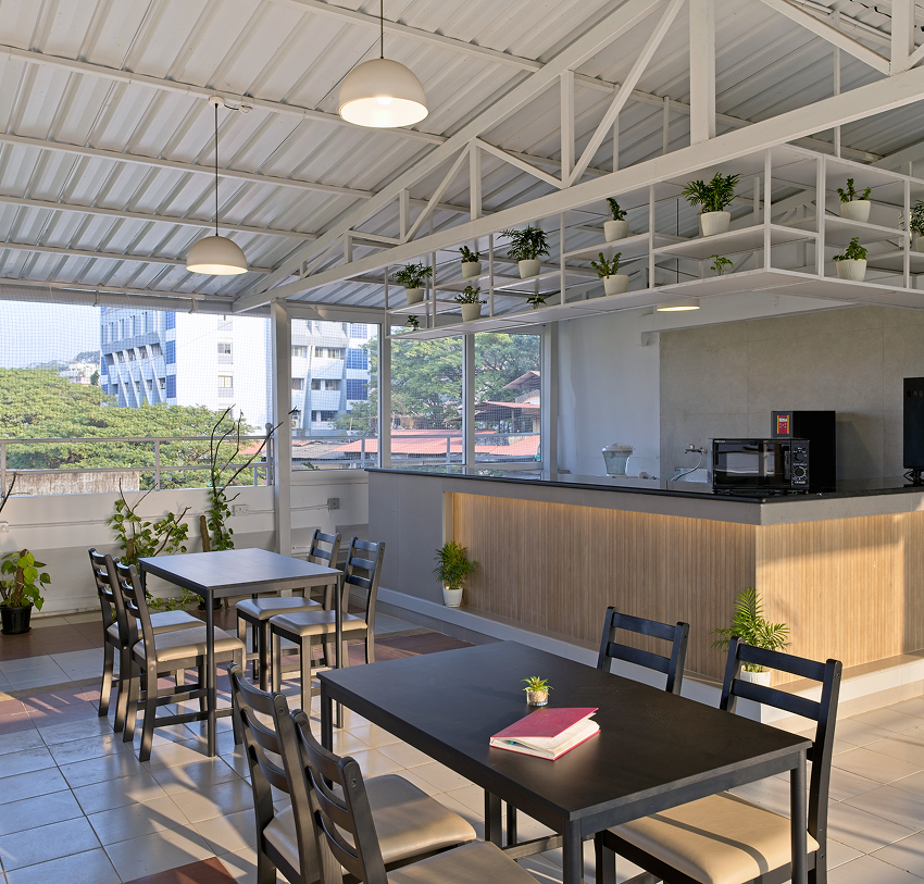 Bright pantry area at Kowo WorkSpace featuring black dining tables and chairs, a modern wooden counter with appliances, hanging lights, indoor plants, and large windows overlooking greenery and nearby buildings.