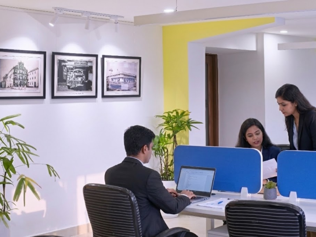 Coworking office scene with professionals collaborating at white desks with blue privacy panels, one person working on a laptop while two colleagues review documents, surrounded by indoor plants and framed wall art.
